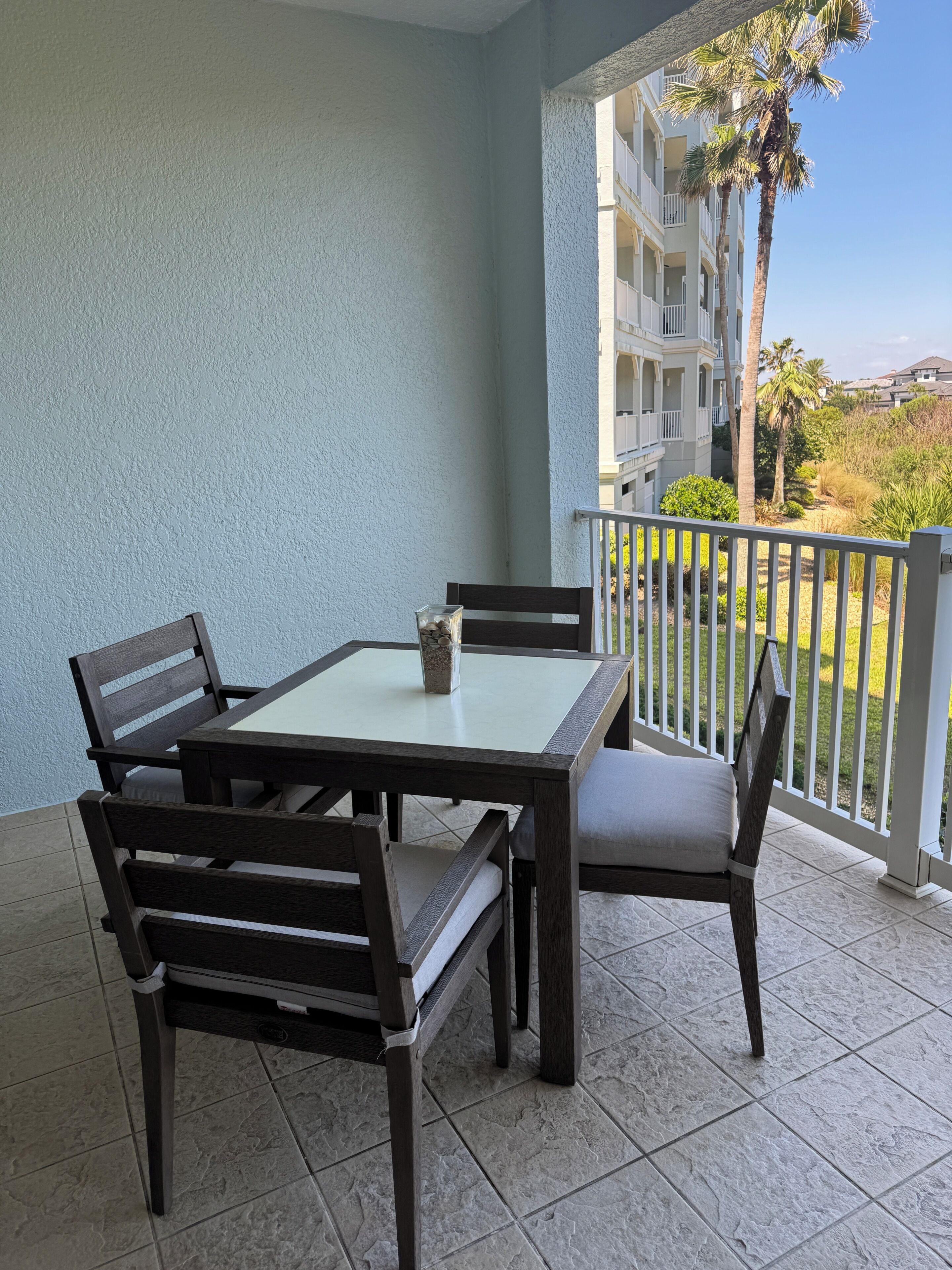 A balcony with a table and chairs, a view of a building, and a palm tree.