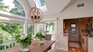 A dining area with a wooden table, white chairs, and a chandelier. There is a view of greenery outside through large windows.