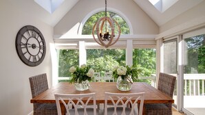 A dining area with a wooden table, white wicker chairs, a large wall clock, and a chandelier.