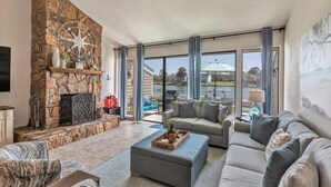 A spacious living room with a stone fireplace, a large window with curtains, a sofa, and a coffee table. - Lake Conroe WATERFRONT Updated 3BR Condo with Pools, Paddle Boards & Views (Montgomery)