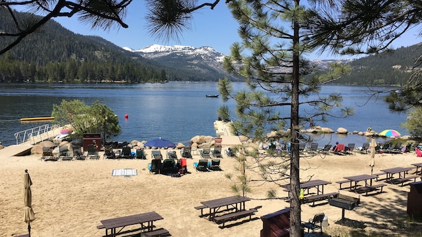 A lakeside beach with picnic tables and chairs, a barbecue grill, and a view of a mountain range.