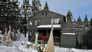 A two-story wooden cabin with a red door, surrounded by snow-covered ground and trees.