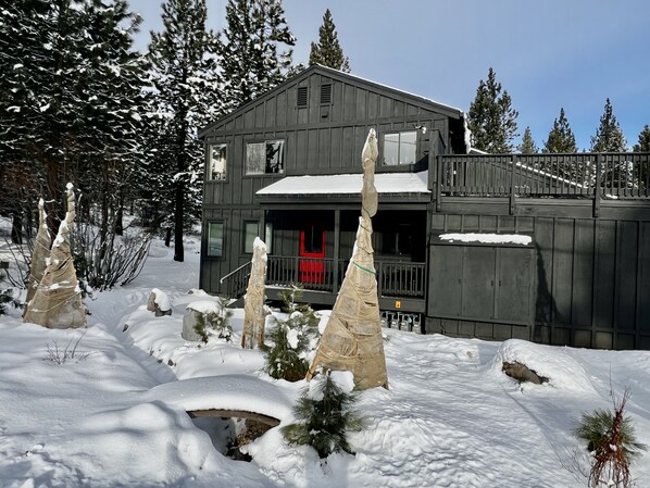 Una cabaña de madera de dos pisos con una puerta roja, rodeada de nieve y árboles.