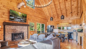 A cozy cabin interior with a stone fireplace, a large gray sofa, and a kitchen area. - Mtn View Cabin 5 min to Downtown Blue Ridge w/Hot Tub and Firepit, Pet Friendly! (Blue Ridge)