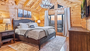 A wooden cabin bedroom with a bed, nightstands, a chandelier, and a large window. - Mtn View Cabin 5 min to Downtown Blue Ridge w/Hot Tub and Firepit, Pet Friendly! (Blue Ridge)