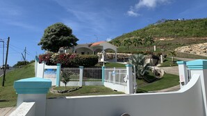 A gated property with a white building, a large tree, and a hill in the background. - Gated "Sea Breeze" House Overlooking the Caribbean (Patillas)
