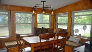 A wooden cabin interior with a dining table and chairs, large windows with a view of a lake, and a ceiling light fixture.