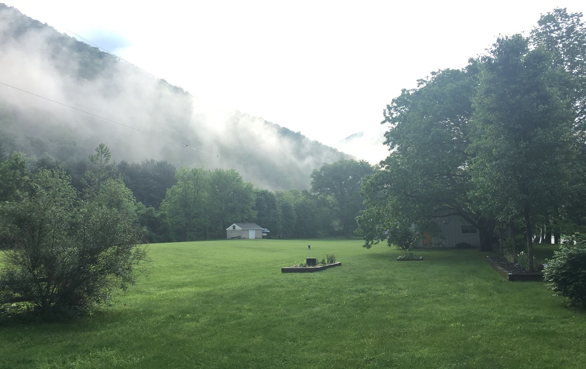 A grassy field with a small house and a boat, surrounded by trees and mountains.