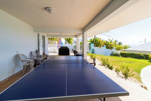 Mesa de ping pong cubierta al aire libre con sillas, una parrilla y vistas a una zona residencial con valla blanca y vegetación.