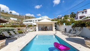En swimmingpool med solstolar, en rosa uppblåsbar badring och en byggnad med en balkong i bakgrunden.