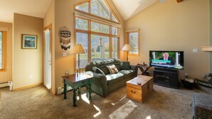 A living room with a high ceiling, large windows, a television, a sofa, a coffee table, and a desk with lamps.