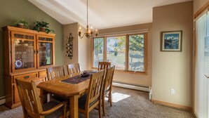 A dining room with a wooden table and chairs, a cabinet with glass doors, a chandelier, and a large window with a view of trees and a lake.