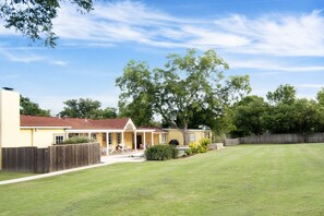 A two-story house with a covered porch, a fenced yard, and a large tree. - Sunshine Haus, Sleeps up to 14,  Hot Tub, Pool, Walk to Main Street, 2 firepls (Fredericksburg)