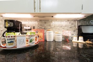 Een keukenblad met een koffiezetapparaat, theemokken en koffiemokken, een boek, zout- en pepervaatjes, en een decoratieve spin.