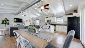 Un vaste espace de vie comprenant une table à manger en bois, des chaises blanches, un canapé gris et un ventilateur de plafond.
