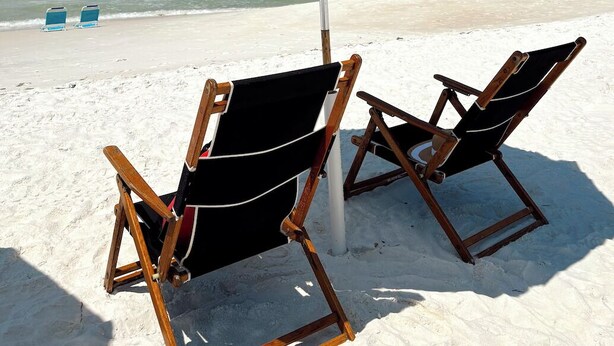 A beach scene with a striped beach umbrella, two wooden beach chairs, and a clear view of the ocean.