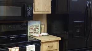Une cuisine avec des meubles en bois, un micro-ondes noir, un réfrigérateur noir et un panneau indiquant : « COUNTRY ROADS, l’ambiance de la campagne, chez moi, là où j’ai ma place ».