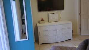 Une pièce avec un grand miroir, un téléviseur fixé au mur, un meuble en osier et une porte.