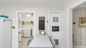 A clean, white-walled laundry room with a table and chairs, a hanging laundry basket, and a door with a 'MENU' sign.