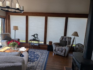 Uma sala de estar acolhedora com lareira, uma mesa de centro com um vaso de flores e uma janela grande com persianas.