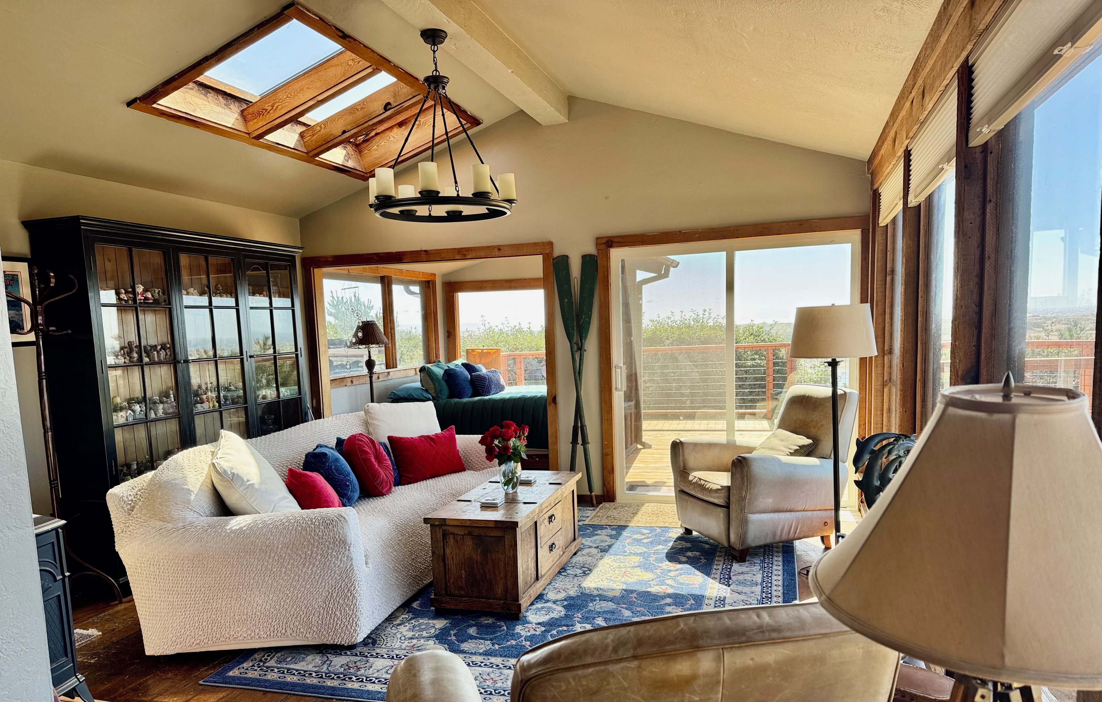 Un salon spacieux avec un plafond haut, des fenêtres de toit, un grand canapé agrémenté de coussins colorés, une table basse en bois et une vue sur une terrasse à travers de grandes fenêtres.