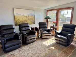 A room with four black leather chairs arranged around a small table, a large painting on the wall, and a window with a view of trees and a house.