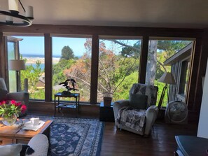 A room with large windows, a chair, a table with a vase of flowers, and a fan.