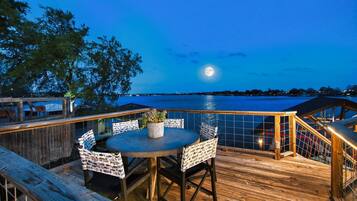 A wooden deck with a round table and chairs, overlooking a body of water with a moon in the sky.