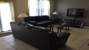 Una sala de estar moderna con un sofá de cuero negro, dos mesitas de noche con lámparas, una mesa de centro de vidrio y un televisor de pantalla plana sobre un mueble.