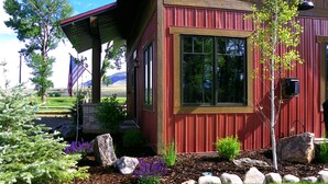 A wooden house with a red exterior and a covered porch. There is a garden with rocks, shrubs, and a tree.