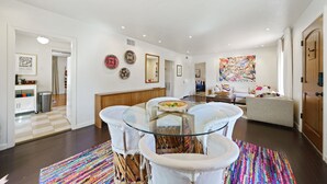 Un vaste espace de vie comprenant un coin repas avec une table à plateau en verre, entouré de chaises blanches à coussins, un tapis coloré et une cuisine visible au fond.