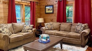 A cozy cabin interior with two leather sofas, a wooden coffee table, and red curtains.