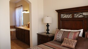 A bedroom with a wooden headboard, a bedside lamp, patterned bedding, and a view into the bathroom with a shower curtain.