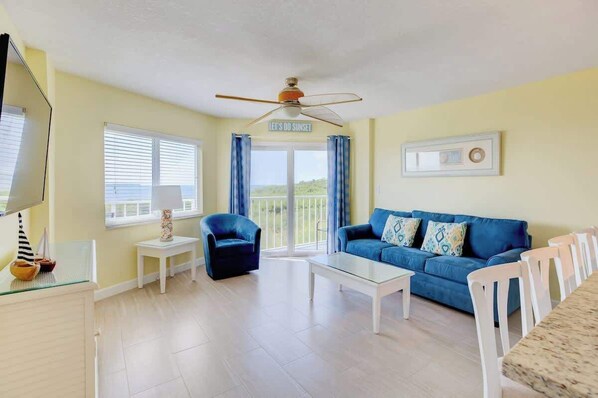 A living room with a blue sofa, white coffee table, and a ceiling fan. There is a window with curtains and a sign that reads "LET'S DO SUNSET".