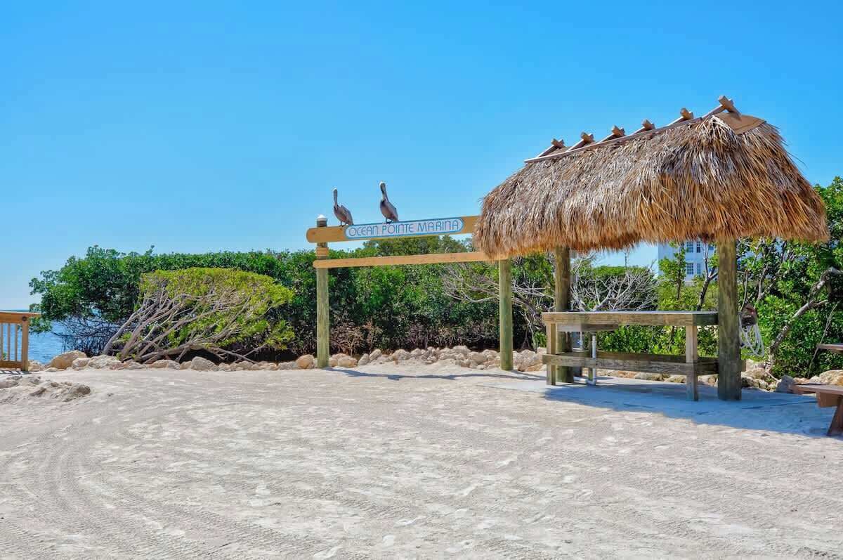 Une entrée de plage avec un panneau indiquant « Ocean Pointe Marina », une structure à toit de chaume et un banc en bois.