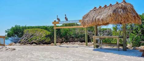 Une entrée de plage avec un panneau indiquant « Ocean Pointe Marina », une structure à toit de chaume et un banc en bois.
