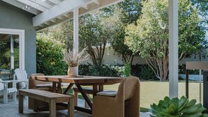 An outdoor patio with a wooden table and wicker chairs, surrounded by greenery and a white pergola.