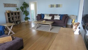Ein Wohnzimmer mit Holzboden, einem braunen Sofa mit Kissen, einem Couchtisch, einem Bücherregal und einer Zimmerpflanze.