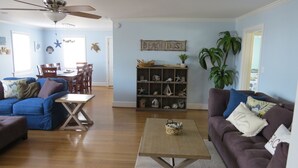 Ein Wohnzimmer mit blauer Wand, Holzboden, Deckenventilator, Essbereich mit Tisch und Stühlen sowie ein Bücherregal mit maritimer Dekoration.