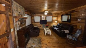 A cozy cabin interior with a stone fireplace, wooden walls, a ceiling fan, and a television.