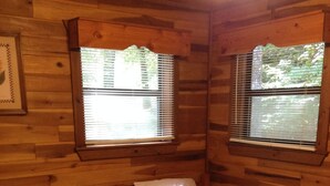 A cabin with two wooden beds, each with a pillow and a quilted blanket. There are two windows with blinds, and a framed picture on the wall.