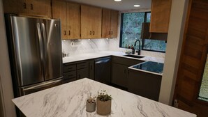 Una cocina moderna con refrigerador de acero inoxidable, armarios de madera, encimera de mármol y ventana con vista a árboles.