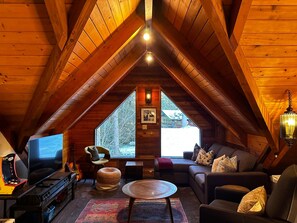A cozy attic room with a sloped ceiling, a large window, a television, a sofa, and a small table.