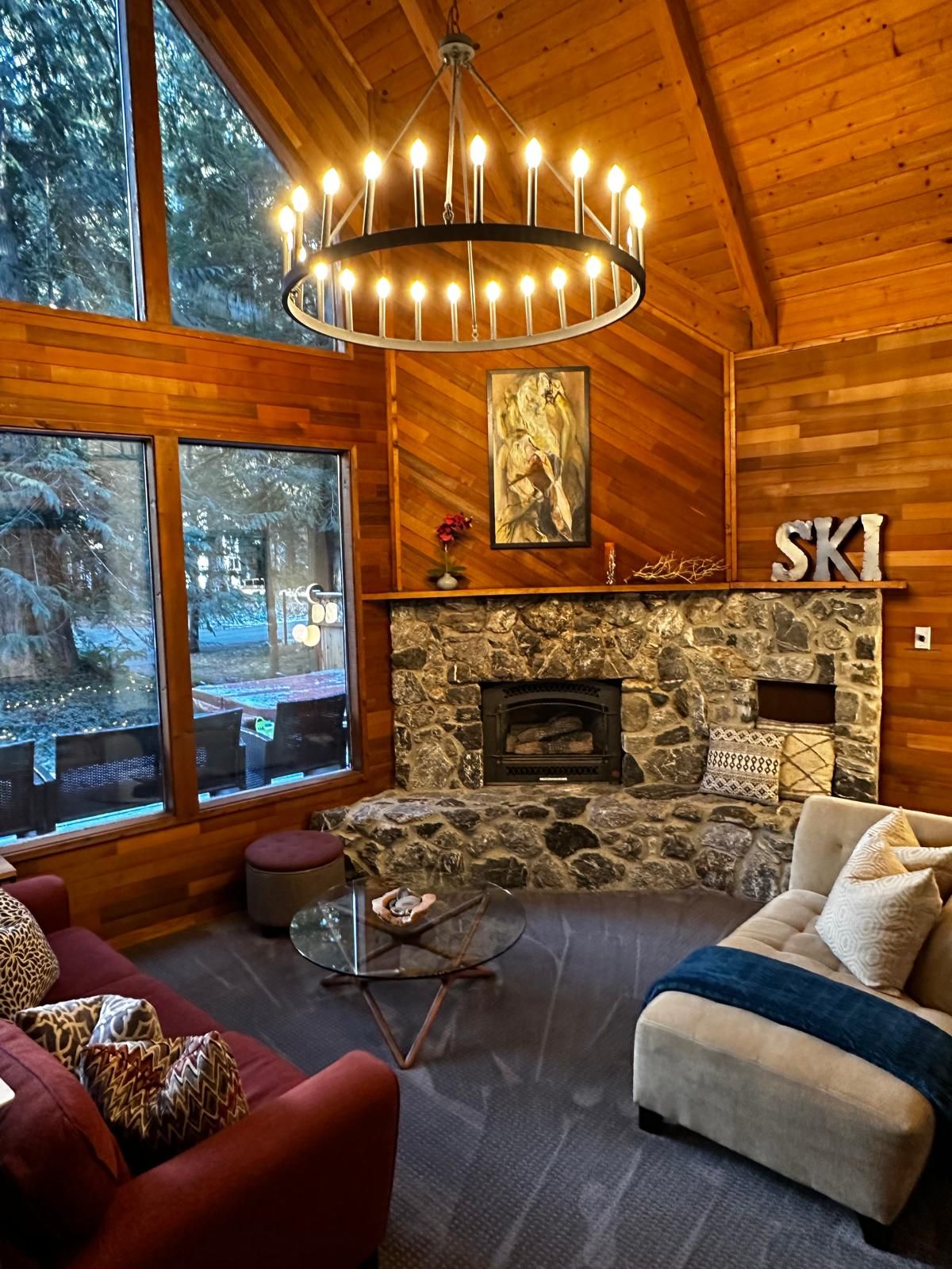 A cozy interior with a stone fireplace, wooden walls, and a large window overlooking a snowy landscape.