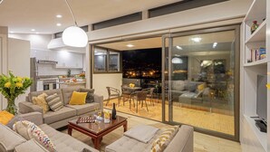Uma sala de estar moderna com sofá, mesa de centro e área de jantar com cadeiras e mesa. Uma cozinha pode ser vista ao fundo, e uma grande porta de vidro deslizante dá acesso a uma varanda com vista para a cidade à noite.