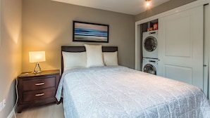 A bedroom with a bed, a nightstand, a lamp, a painting, and a washing machine.