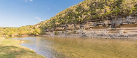 A river with clear water, surrounded by grassy banks and a rocky cliff with vegetation.