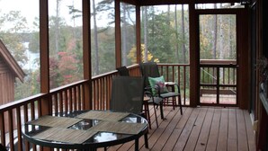 Une terrasse en bois couverte, avec une table ronde en verre et des chaises, offrant une vue sur les arbres et un bâtiment en arrière-plan.