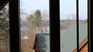 Une vue à travers la fenêtre d’une cabane dont le toit est recouvert de végétation, entourée d’arbres et bordée par une étendue d’eau.