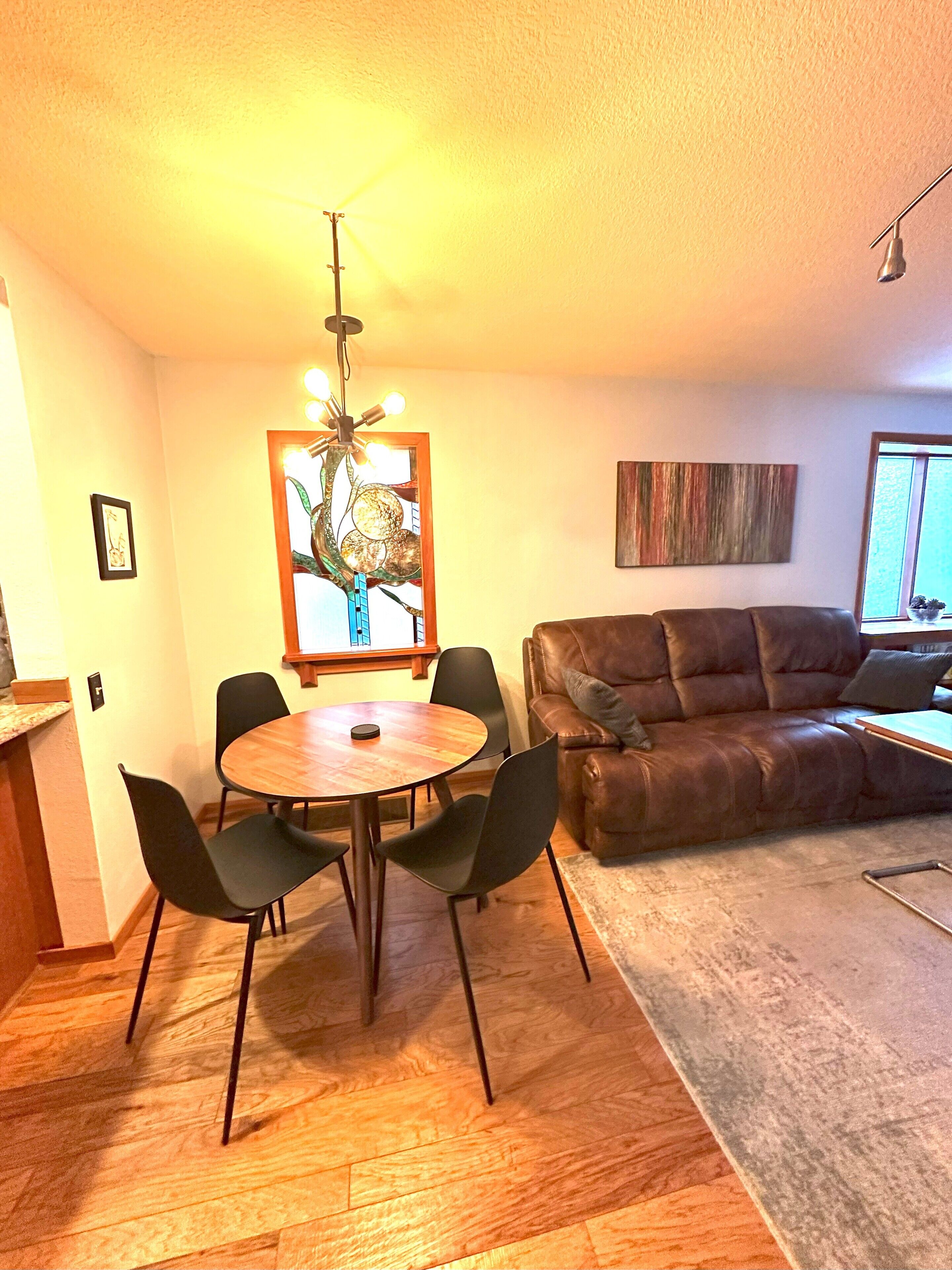 A dining area with a round table and chairs, a brown leather sofa, a decorative wall piece, and a window with a view.